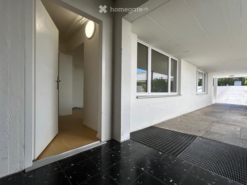 A modern, minimalist hallway with black tiled flooring, white walls, and large windows allowing natural light. The hallway features a wooden door leading to another room and a small, open area with a concrete floor extending outside.