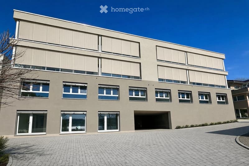 Modern two-story building with large windows, a flat roof, and a paved driveway. Features include a garage entrance and multiple floor-to-ceiling windows.