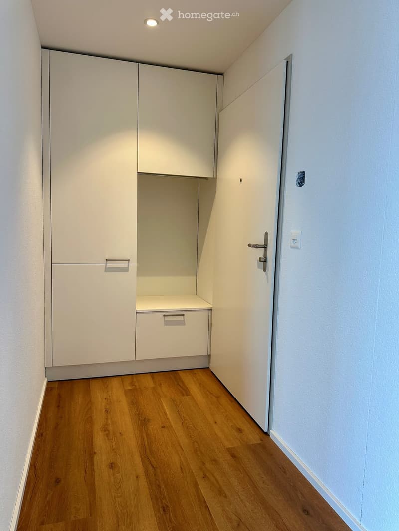 Empty room with white walls, wooden floor, white cabinets, and a door.