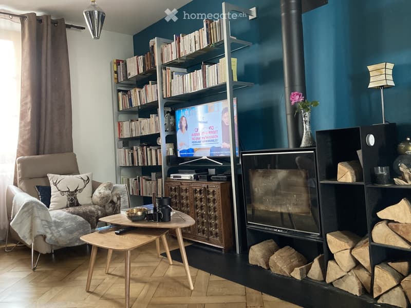 Modern living room with a fireplace, a sofa with a fur throw, a wooden coffee table, a bookshelf filled with books, and a television. The room features a dark blue accent wall and a wooden floor.