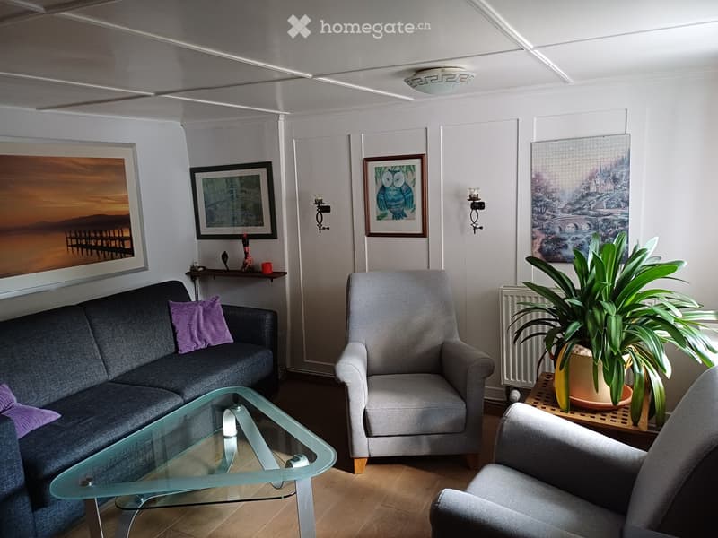 Living room with a grey sofa, armchair, glass coffee table, potted plant, and several picture frames on the wall.