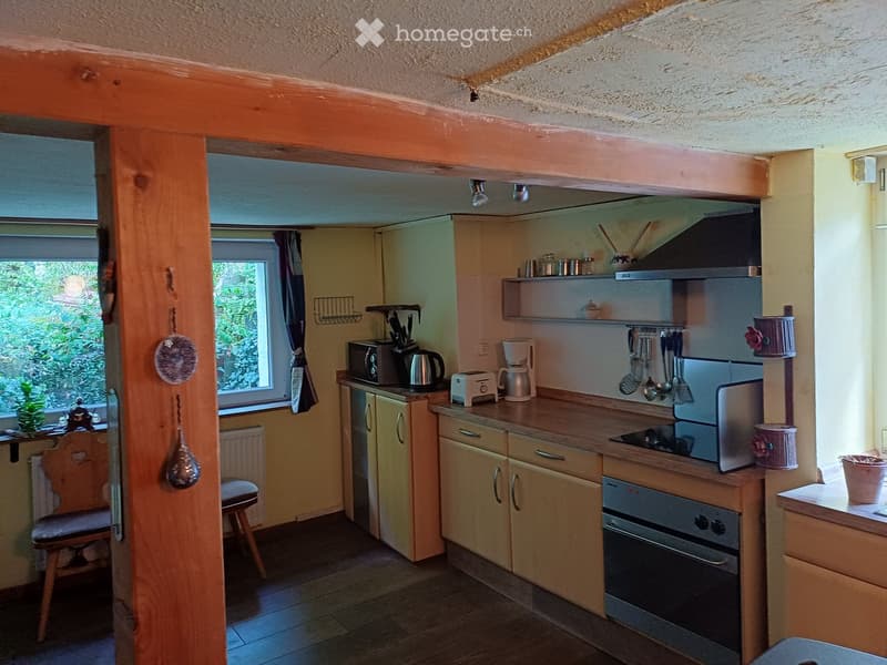 Cozy kitchen with wooden cabinets, stainless steel appliances, and a small wooden stool near the window. The space features a microwave, coffee maker, toaster, and various kitchen utensils hanging from the wall.