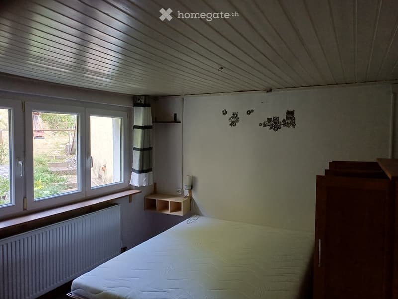 Bedroom with a single bed, wooden wardrobe, small shelves, and a window with a curtain. The room has a slanted ceiling and a radiator under the window.