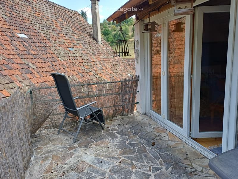 Balcony with stone tile flooring, a black folding chair, a wooden fence, and a view of a tiled roof and greenery.