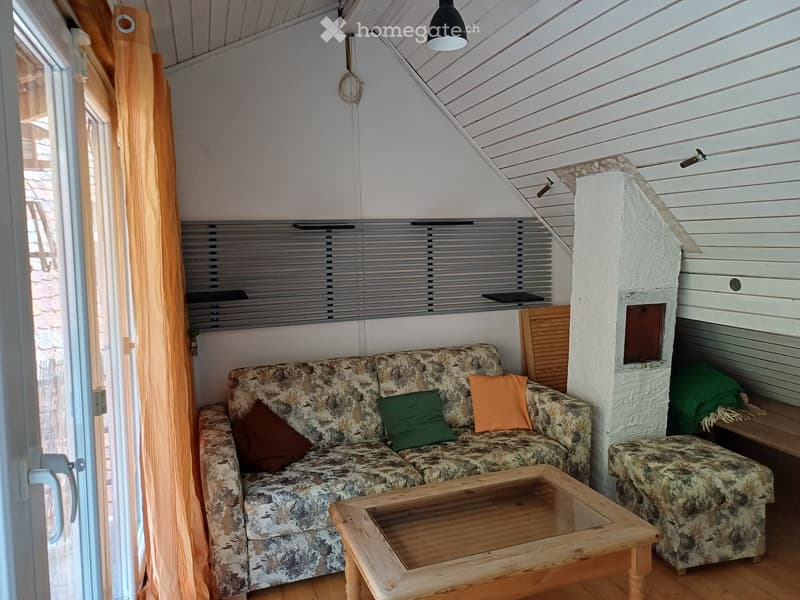 Cozy living room with a patterned sofa, wooden coffee table, armchair, and colorful pillows. Features wooden flooring, large windows with blinds, and a slanted ceiling with exposed beams.
