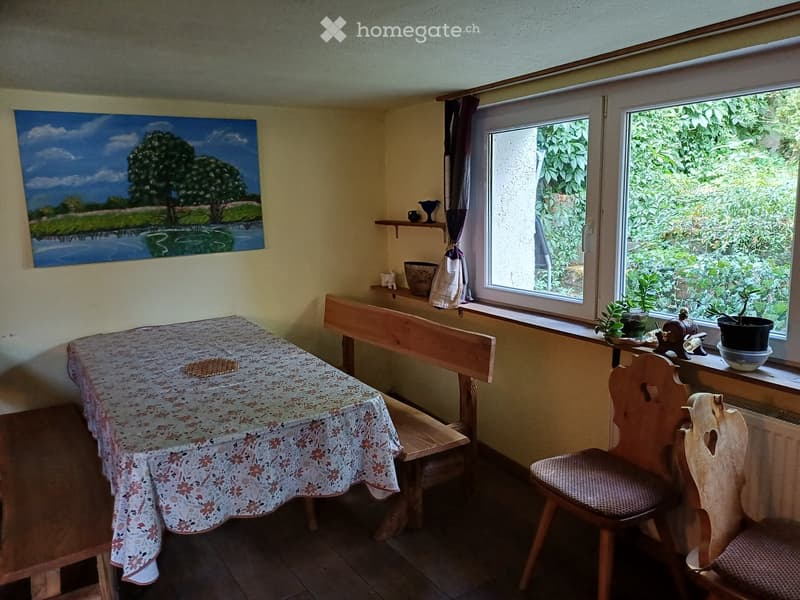 Cozy dining area with wooden bench seating, floral tablecloth, wooden chairs, and a painting on the wall. Windows offer a view of greenery, with small potted plants on the windowsill.