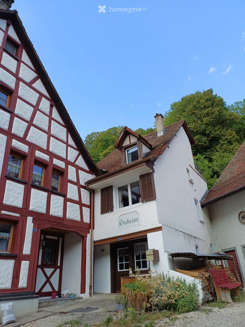 Traditional half-timbered building with white plaster and red wooden framing, featuring multiple windows, a central entrance, and a small garden area with plants and a bench.