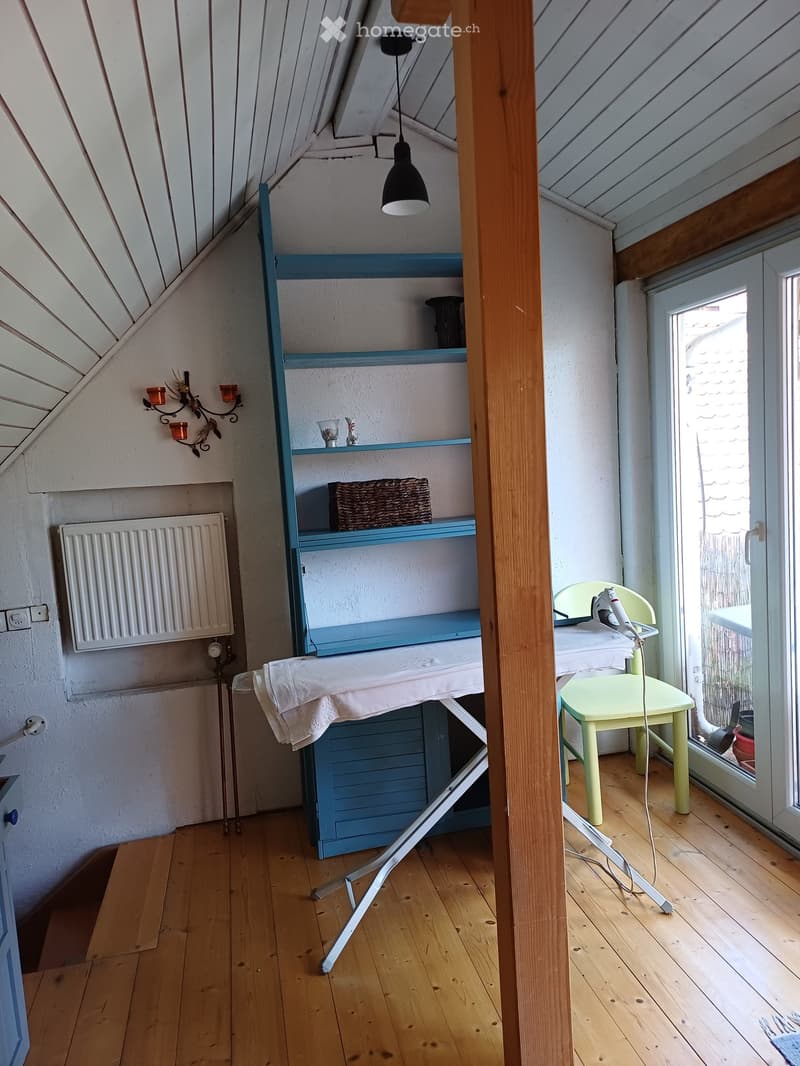 A small attic room with wooden flooring, a slanted ceiling, a light green chair, an ironing board, a blue shelf, and a radiator.