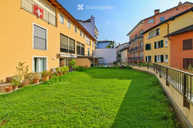 A well-maintained garden area with green grass, potted plants, and surrounding residential buildings with balconies and colorful facades.