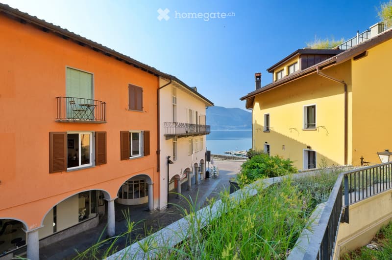 A picturesque street lined with colorful buildings, including orange, yellow, and beige facades, with balconies, shutters, and a view of a lake and mountains in the background.