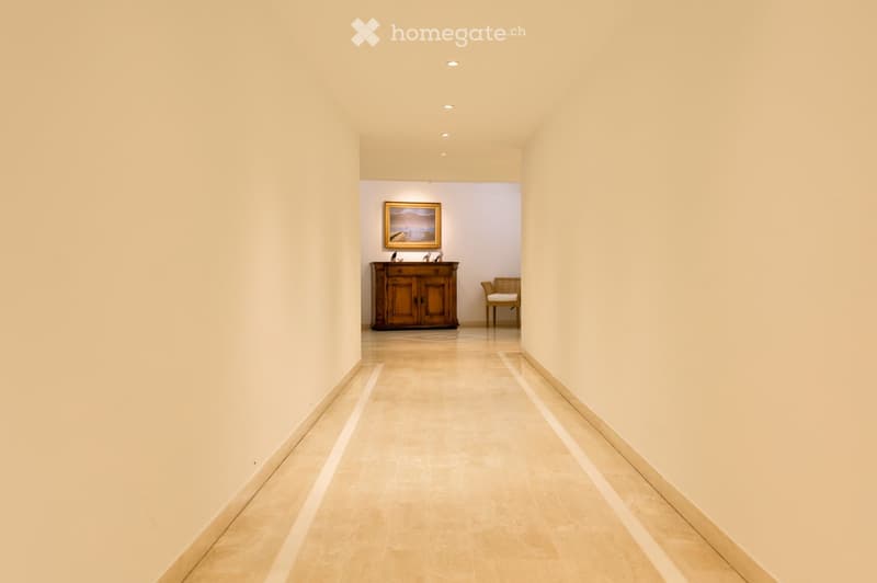 A long, well-lit hallway with beige walls and a polished tile floor, leading to a room with a wooden cabinet, a framed painting, and a wicker chair.