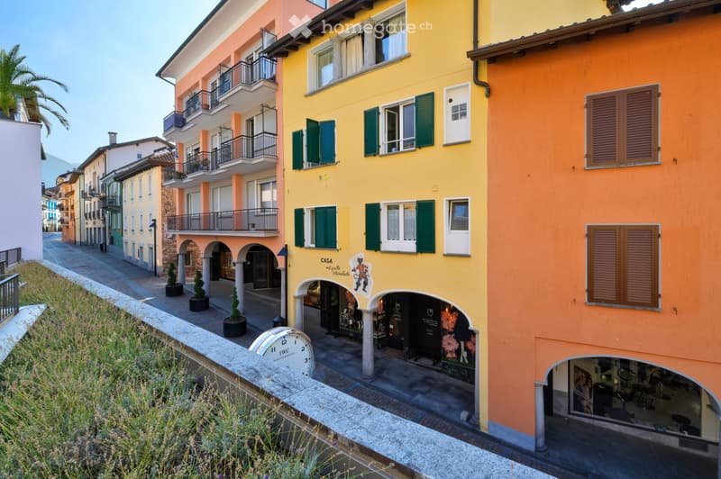 A picturesque street view showcasing colorful multi-story buildings with balconies, green shutters, and arched storefronts. The architecture features a mix of pastel-colored facades and classic European design elements.