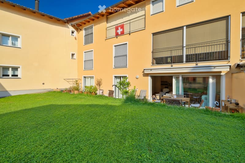 A well-maintained garden area with a green lawn, potted plants, outdoor furniture, and a building with balconies and a Swiss flag.