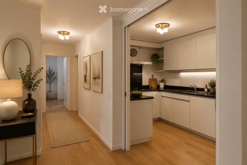 Modern hallway leading to a compact kitchen area with white cabinets, black countertops, and stainless steel appliances. The hallway features a round mirror, framed pictures, and a small table with a lamp and vase.