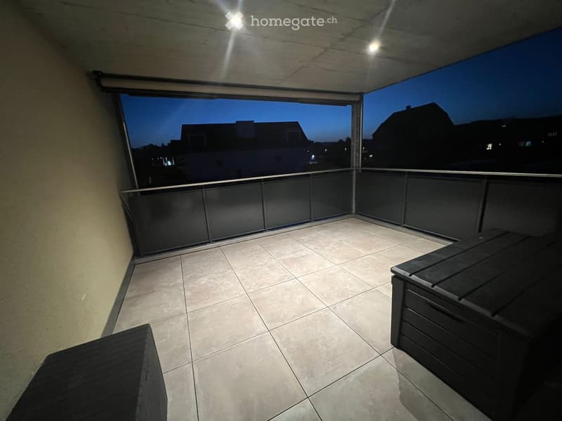 Modern balcony with tiled floor, glass railing, and two dark-colored lounge chairs.