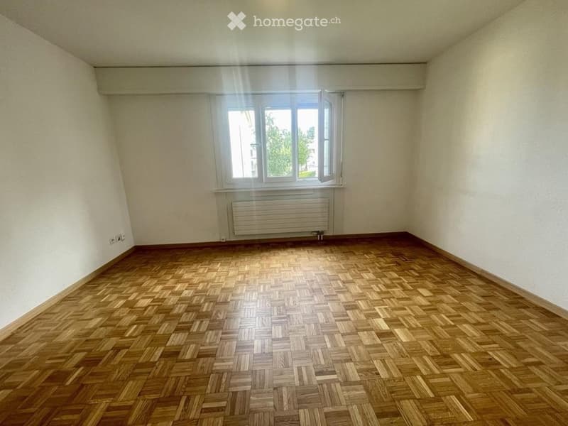 Empty room with wooden parquet flooring, white walls, and a single window with a radiator below it.