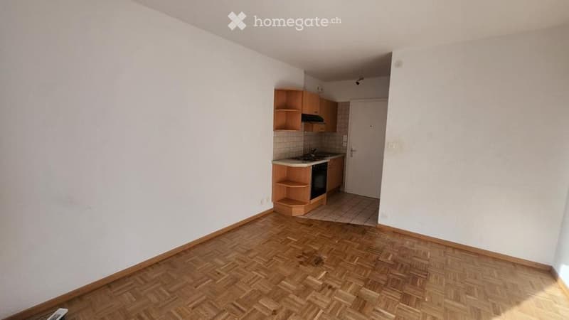 Empty room with wooden parquet flooring and white walls, featuring an open kitchen area with cabinets and appliances.
