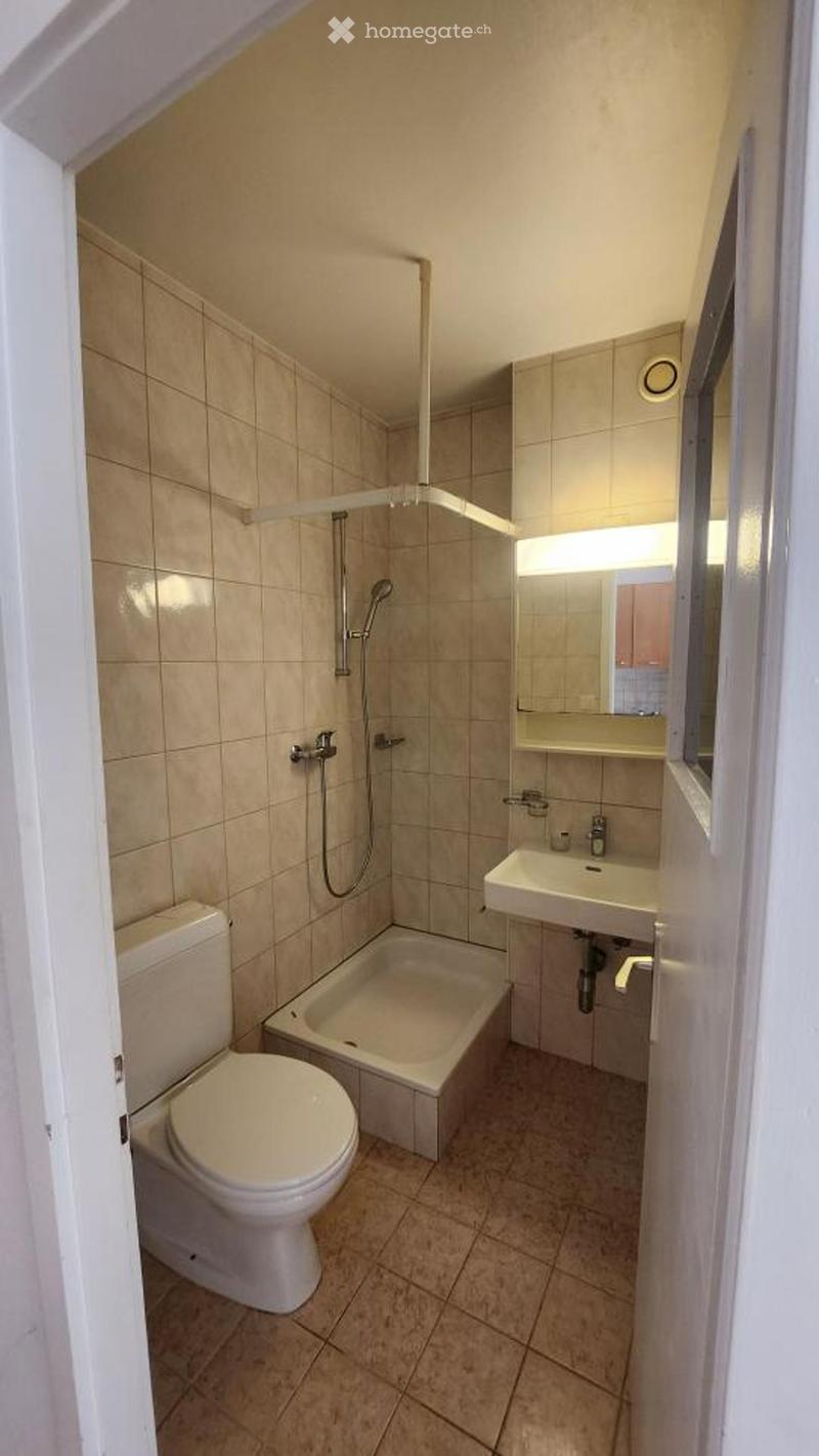 Compact bathroom with beige tiled walls and floor, featuring a toilet, bathtub, sink, and mirror.