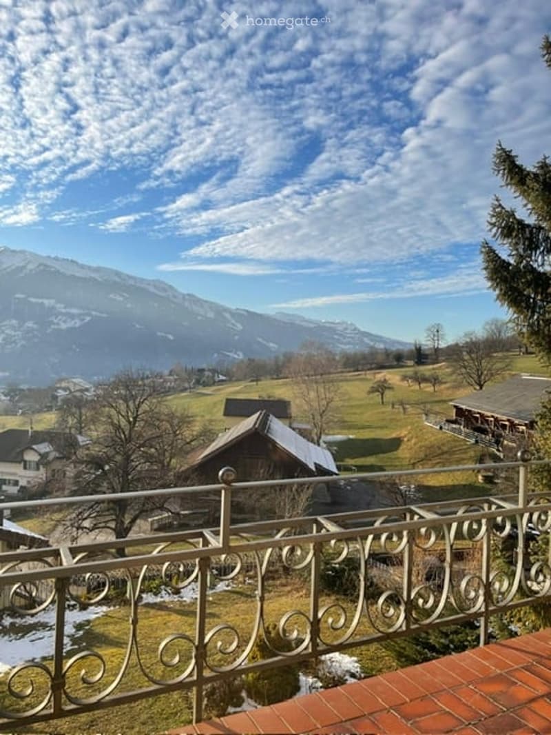 Building in the countryside with a balcony, mountains in the distance, brick-tiled roof, fence, trees