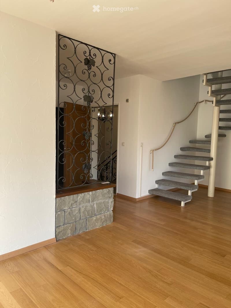 Interior with wood floor, white wall, a spiral staircase with grey steps, black wrought iron decorative wall, an arched brown door, two wall outlets, and a chandelier.