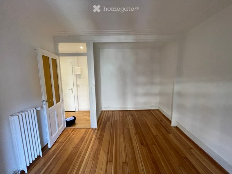 Empty room with wooden flooring, white walls, and a radiator on the left side. A door with glass panels leads to another room.