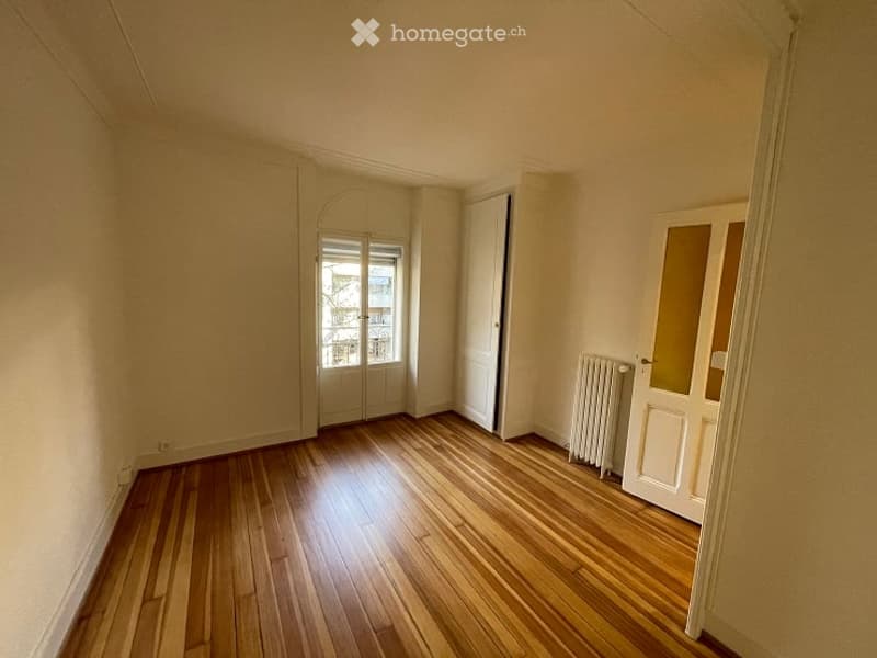 Empty room with wooden flooring, white walls, and a radiator. Features a door with glass panels and a window with frosted glass.