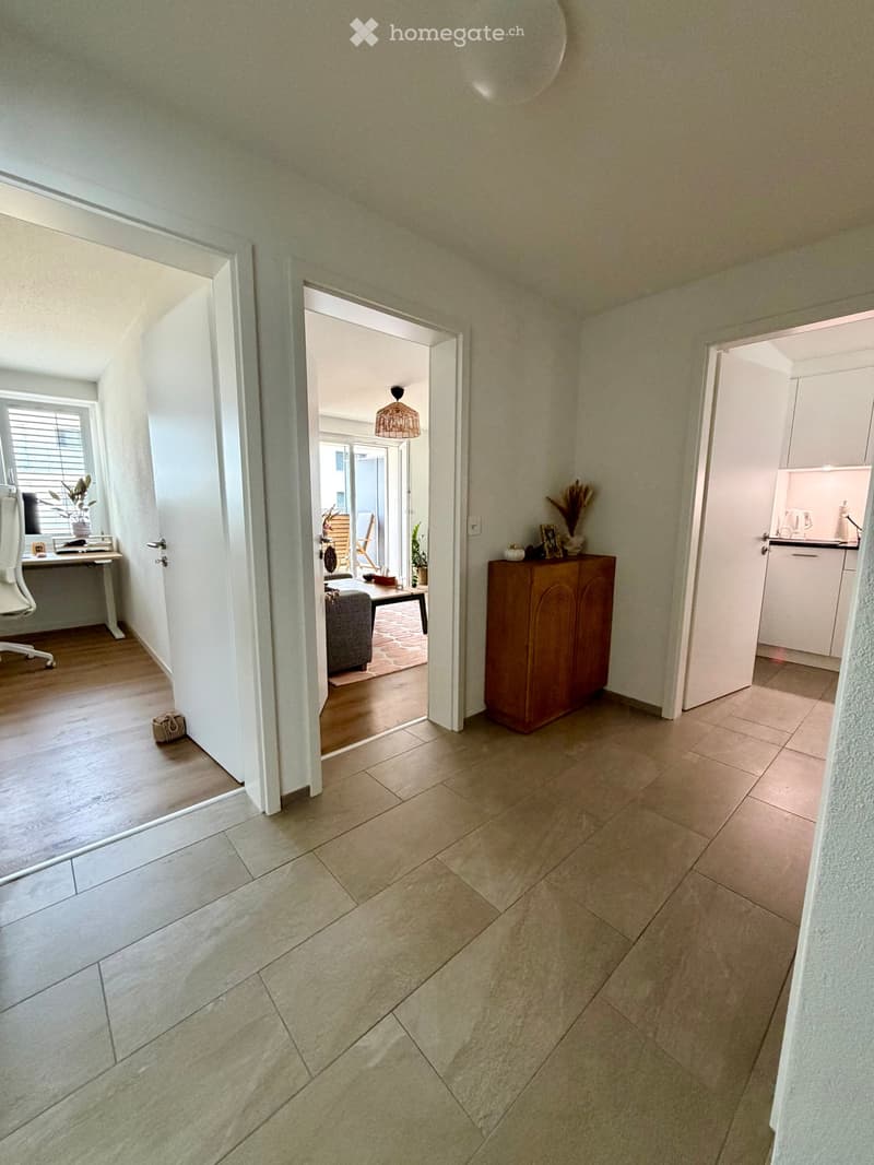 Bright hallway with tiled floor, leading to multiple rooms including a living area, kitchen, and office space. Features include a hanging light fixture, a small wooden cabinet, and a window with blinds.