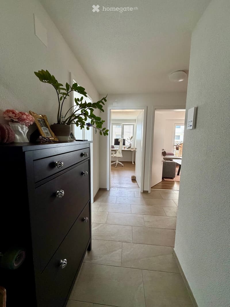 A well-lit hallway with tiled flooring, leading to a room with a desk and chair, and another room with a sofa and coffee table. A dark dresser with decorative items is visible on the left side.