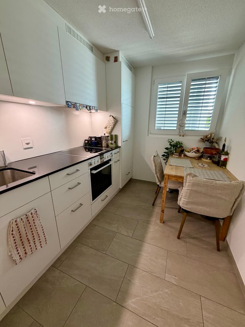 Modern kitchen with white cabinets, stainless steel sink, oven, and a small dining area with a table and chairs. The space features tiled flooring and a window with blinds.