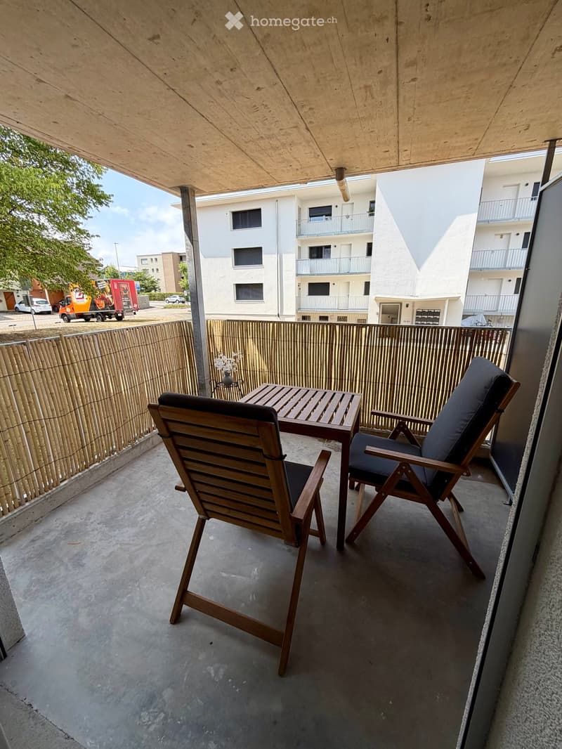 Balcony with wooden table and two chairs, bamboo railing, and view of neighboring buildings.