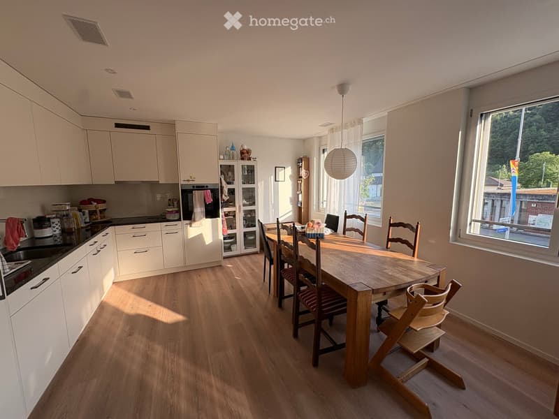 Open-plan kitchen and dining area with light wood flooring, white cabinets, a central dining table with chairs, and a modern kitchen setup including a stove, sink, and dishwasher.