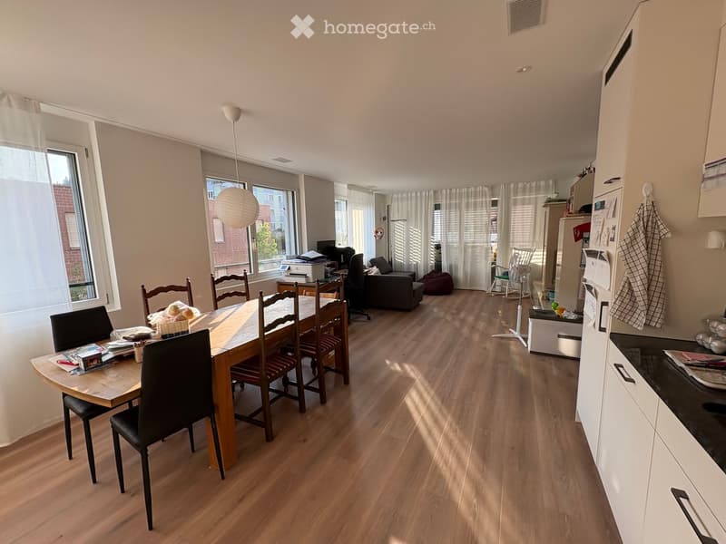 A well-lit, open-plan living and dining area with wooden flooring, large windows, a dining table with chairs, a sofa, a refrigerator, and a fan.
