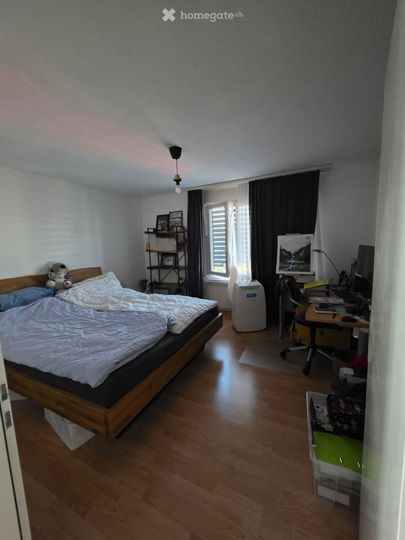 Bedroom with wooden floor, bed with white bedding, desk with chair, shelf with picture frames, air conditioner, and storage box.