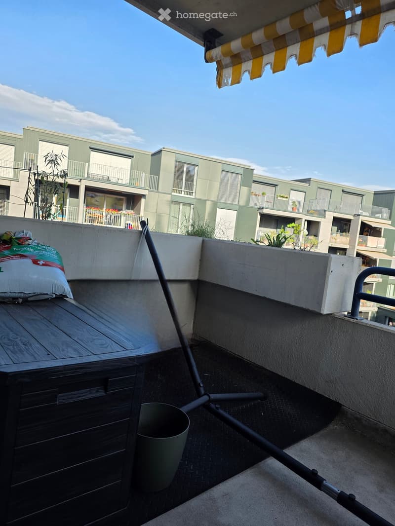 Balcony with wooden table, metal railing, and awning, overlooking a modern apartment complex with balconies and greenery.