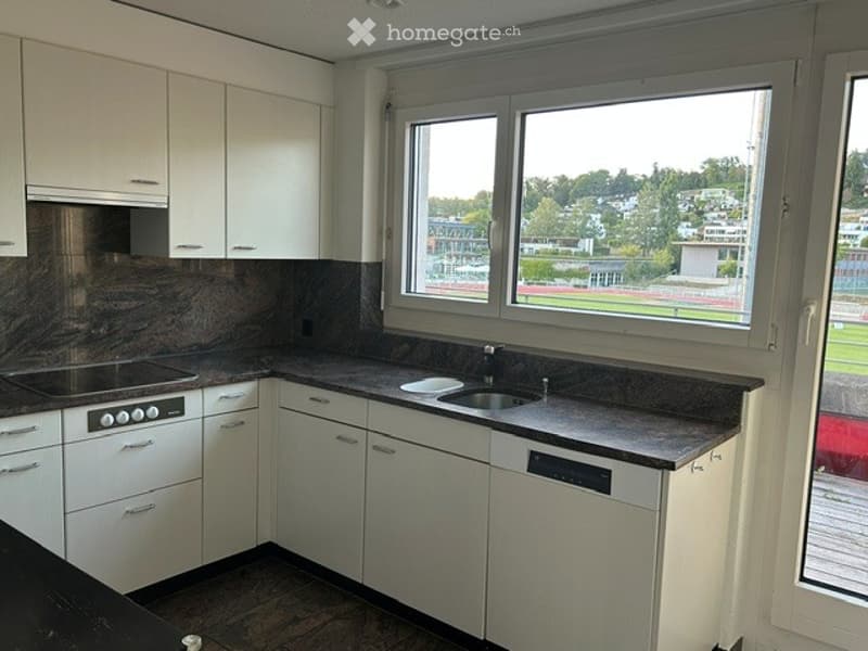 Modern kitchen with white cabinets, granite countertops, and stainless steel appliances. Large windows offer a view of a sports field.