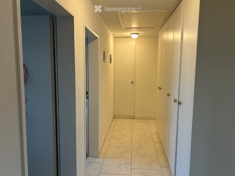 A narrow hallway with light-colored walls, white tiled flooring, and several closed doors on both sides. A single ceiling light illuminates the space.