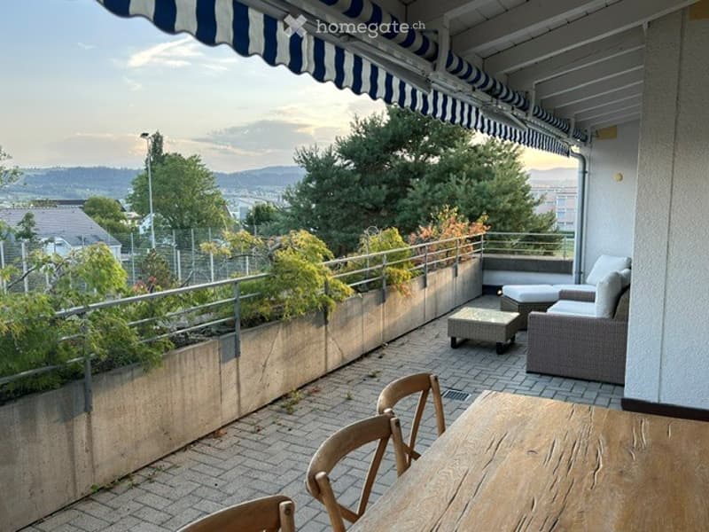 Balcony with outdoor furniture, including a table and chairs, overlooking a scenic view of trees and hills.