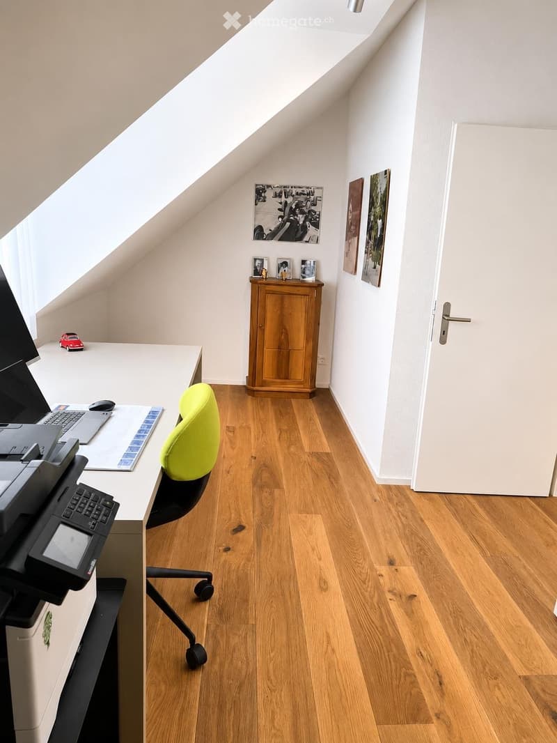 Bright attic room with wooden flooring, slanted ceiling, and large windows. Features a modern desk setup with a chair, printer, laptop, and mouse. A small wooden cabinet holds framed pictures and decorative items. A toy car sits on the windowsill.