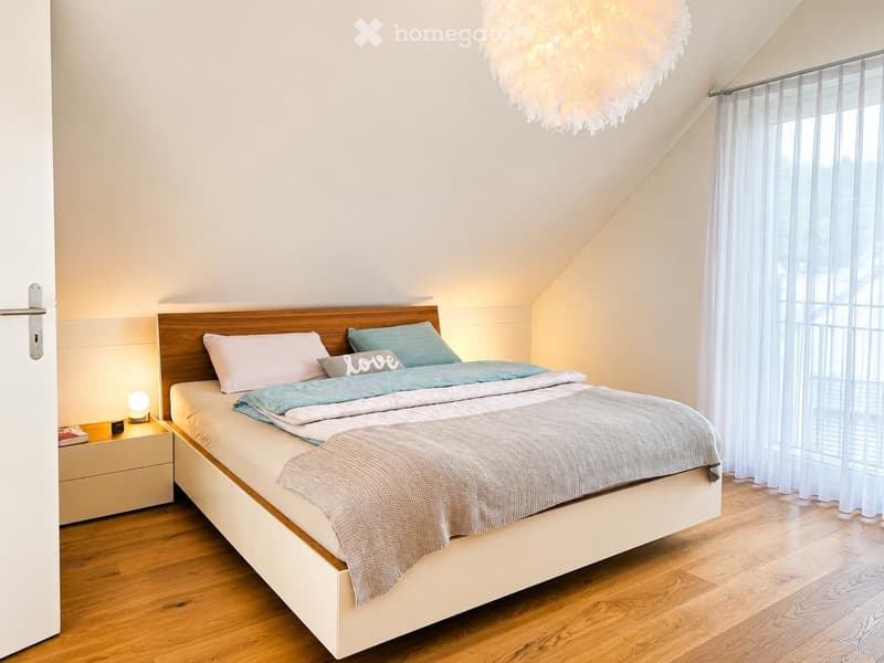 Cozy attic bedroom with wooden flooring, a large bed with pillows and blankets, a nightstand with a lamp, and a decorative chandelier.