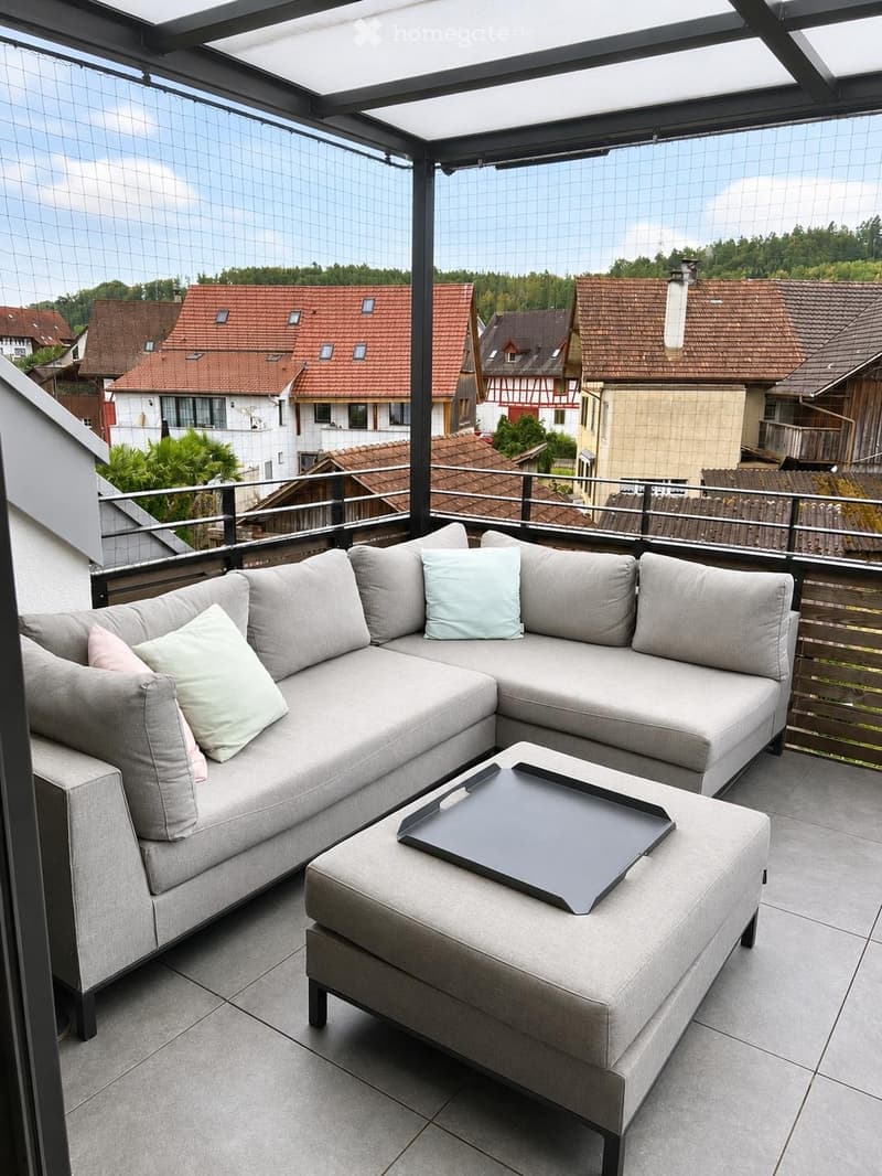 Modern balcony with grey sectional sofa, ottoman, and tray table, featuring a view of the neighborhood.