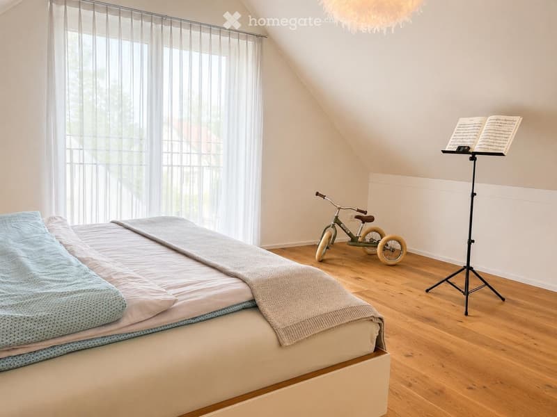 Cozy attic bedroom with a large bed, light blue and white bedding, a small bicycle, and a music stand with sheet music. The room features wooden flooring and large windows with sheer curtains.