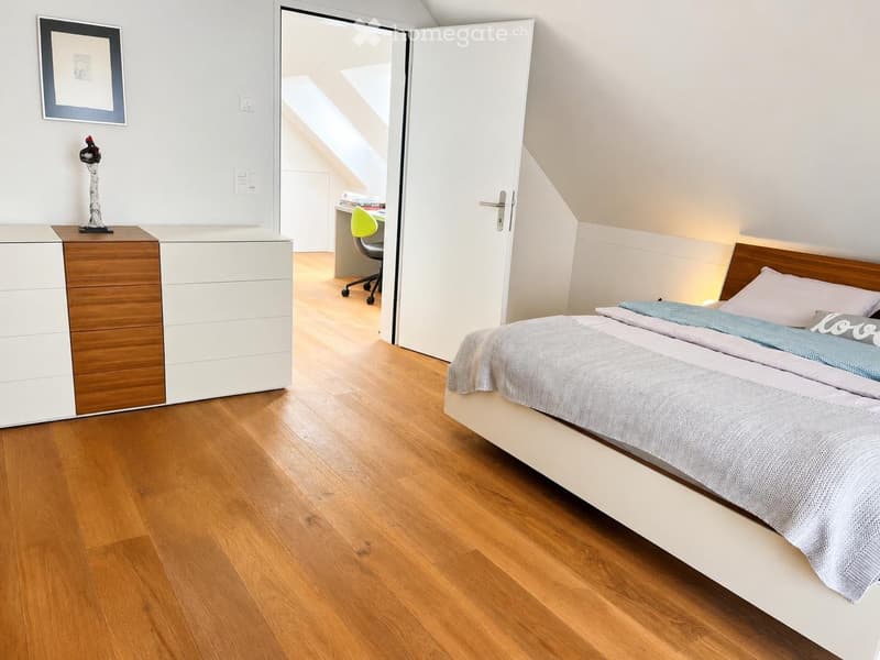Modern bedroom with a wooden bed, nightstand, desk, and chair. The room features light-colored walls and wooden flooring.