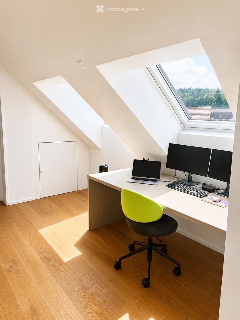 Bright, modern attic office with wooden flooring, large skylights, and a sleek white desk setup including a laptop, dual monitors, keyboard, and a green office chair.