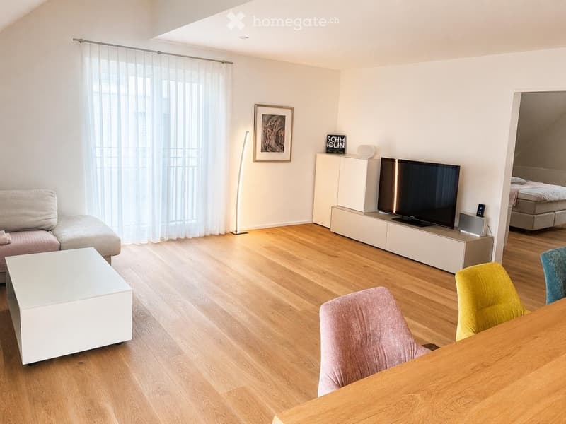 Modern living room with light wooden flooring, white walls, and large windows covered by sheer curtains. Features a grey sofa, white coffee table, colorful chairs, a flat-screen TV on a white cabinet, and a floor lamp.