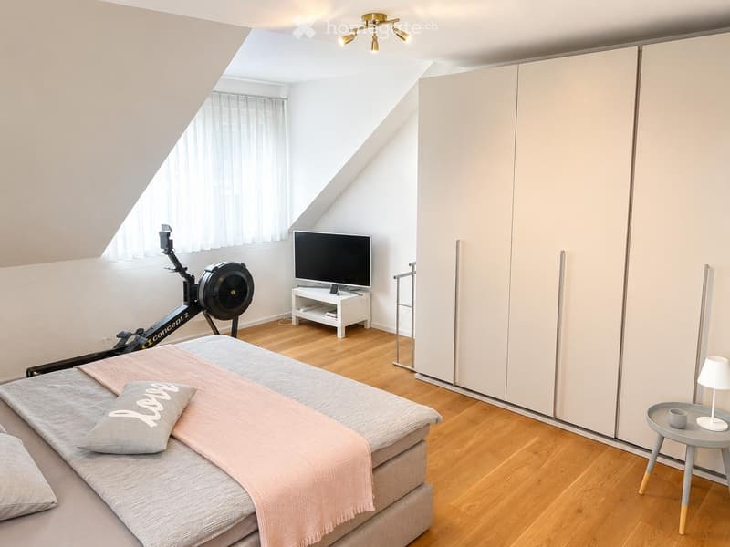Bright bedroom with wooden flooring, a large bed with a pink blanket and grey pillow, a modern wardrobe, a television on a stand, a small round table with a lamp, and an exercise bike near the window.
