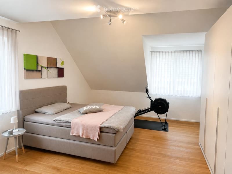 Cozy bedroom with a double bed, nightstand, lamp, exercise bike, and artwork on the wall. The room features wooden flooring and large windows with curtains.
