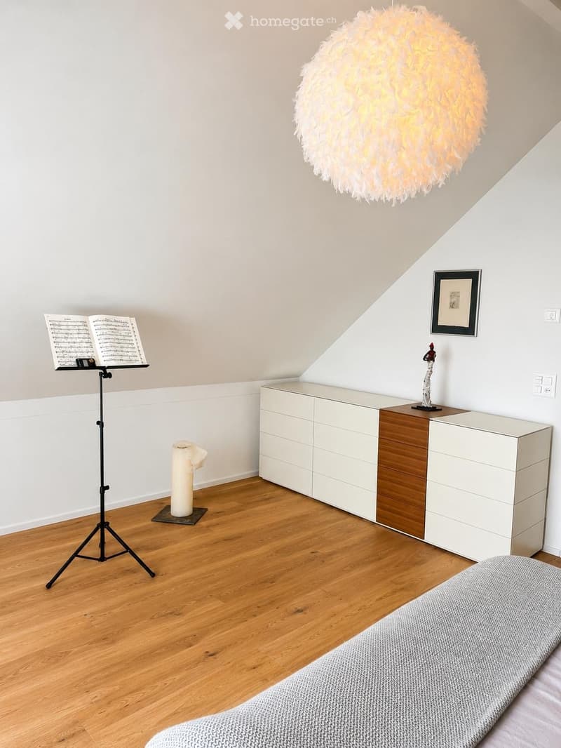 Modern bedroom with wooden flooring, white walls, a large pendant light, a bed, a white cabinet with wooden accents, a cat tree, a music stand with sheet music, and a decorative sculpture on a side table.
