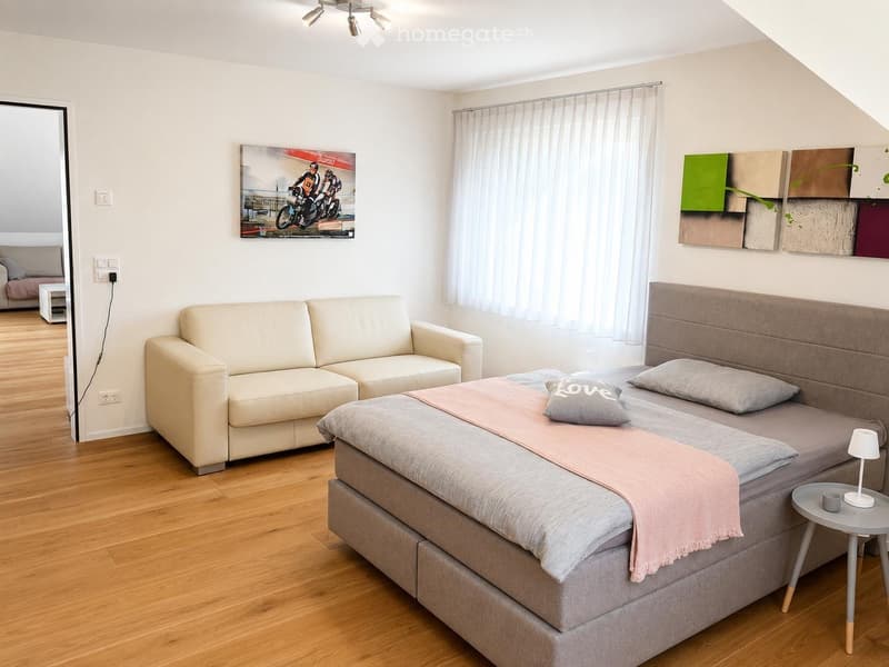 A modern, well-lit room with a sofa, a bed, a nightstand, and a table lamp. The room features wooden flooring, white walls, and large windows with sheer curtains.
