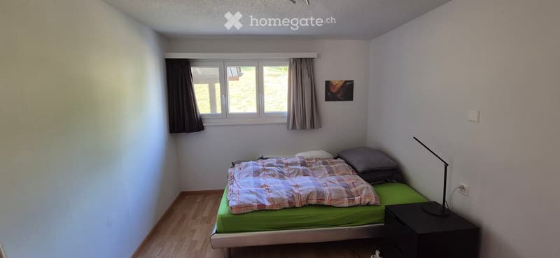 Cozy bedroom with a single bed, green mattress, plaid blanket, nightstand, and a lamp. The room has wooden flooring, white walls, and a window with curtains.