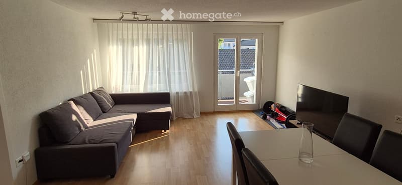 Bright living room with wooden flooring, a grey sectional sofa, a dining table with chairs, and a television. A sliding glass door leads to a balcony.
